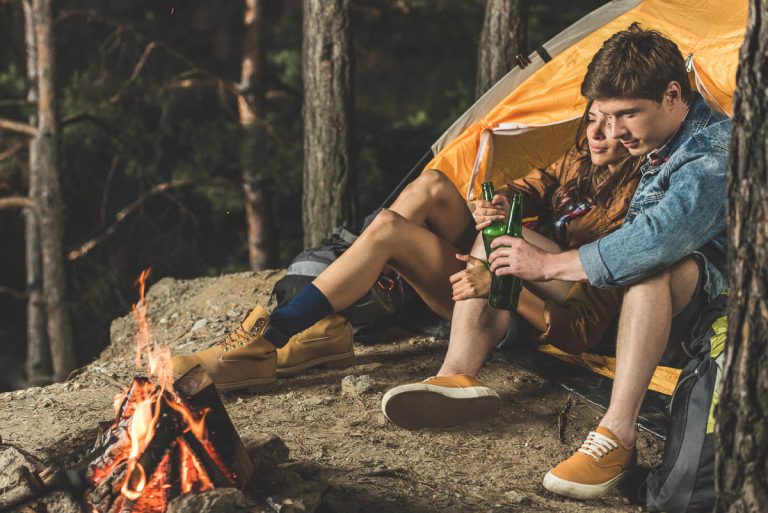 A young couple relaxing at a forest campsite, sitting beside a glowing campfire outside a yellow tent