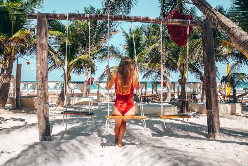 Woman in swimsuit sitting on wooden swing seat on sand and admiring view at beautiful beach resort in tulum, Mexico.
