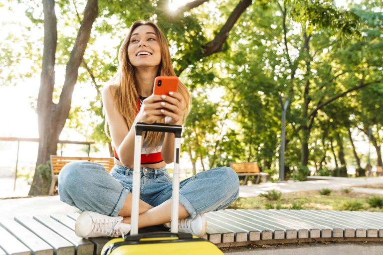 Photo of pretty tourist girl smiling and holding smartphone while sitting with luggage on bench in green park