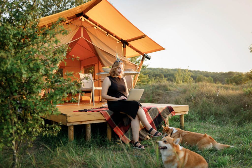 Happy Woman freelancer using a laptop on a cozy glamping tent in a sunny day. Luxury camping tent for outdoor summer holiday and vacation. Lifestyle concept