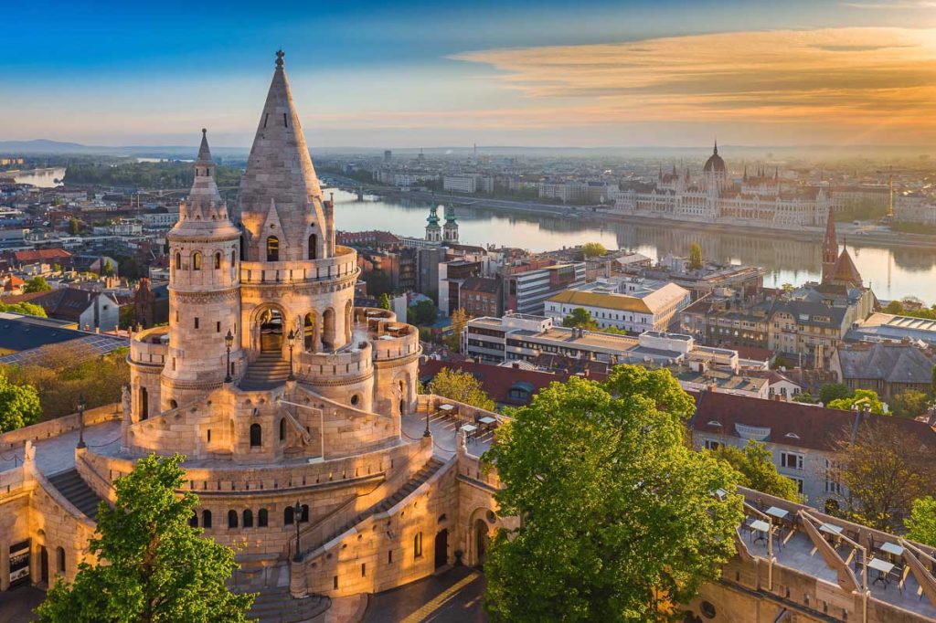 Budapest, Hungary - Beautiful golden summer sunrise with the tower of Fisherman's Bastion and green trees. Parliament of Hungary and River Danube at background. Blue sky.