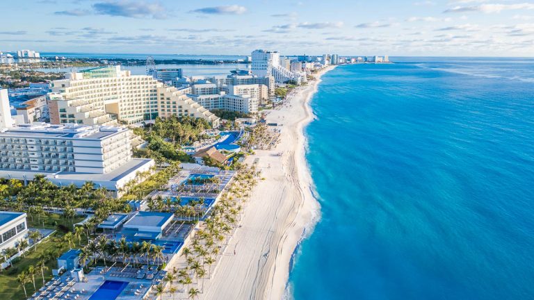 A beautiful beach with a blue ocean and a few buildings in the background. The beach is empty and the sky is cloudy. Aerial Views of Cancun Hotel zone, Quintana Roo Mexico