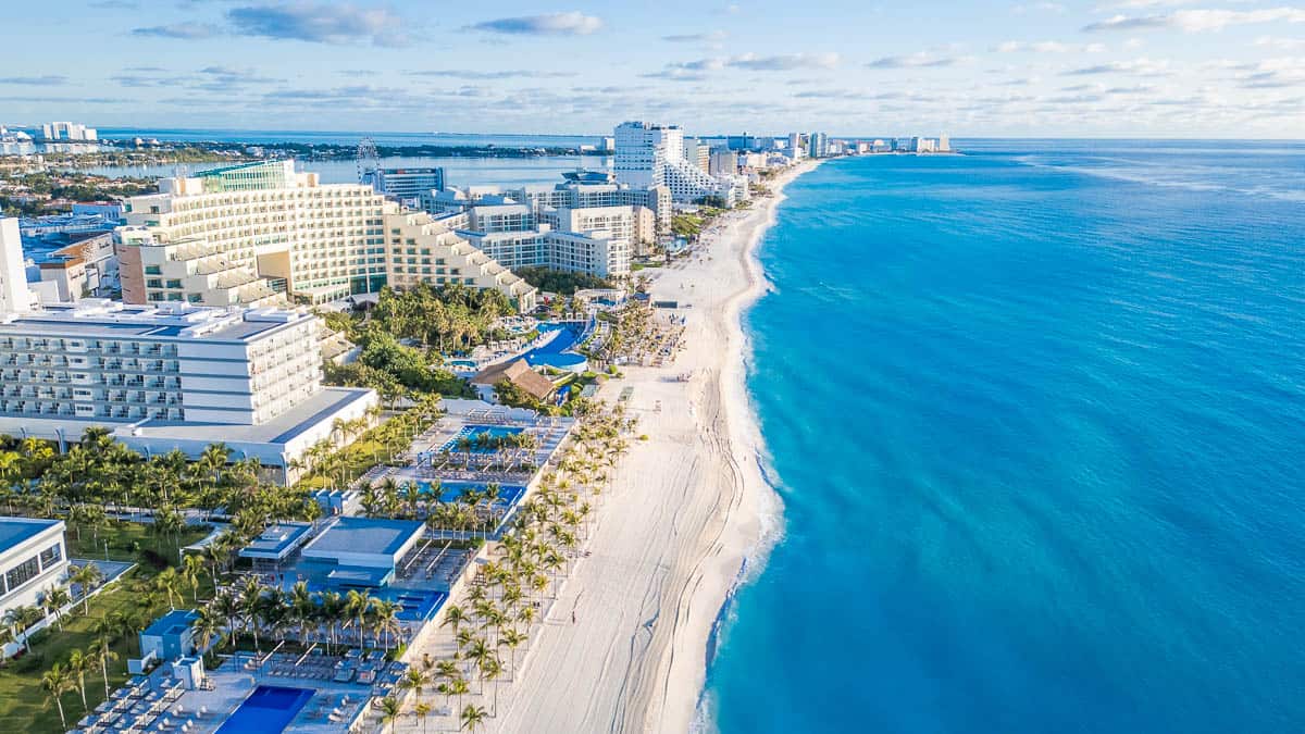 A beautiful beach with a blue ocean and a few buildings in the background. The beach is empty and the sky is cloudy. Aerial Views of Cancun Hotel zone, Quintana Roo Mexico