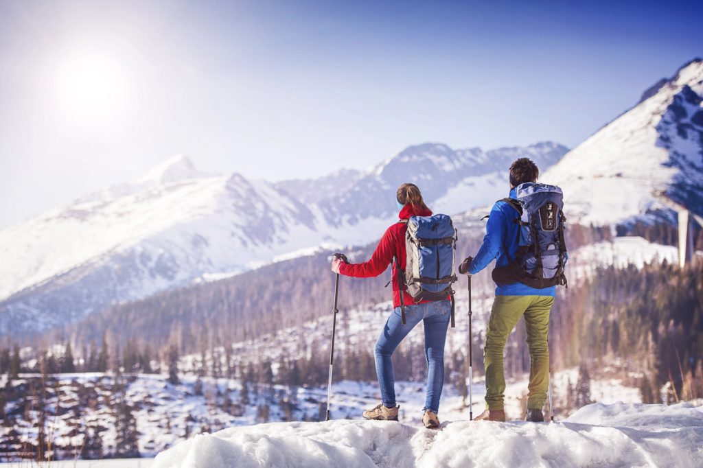 Snowy alpine mountains, two hikers-couple standing on a ridge with trekking poles and backpacks