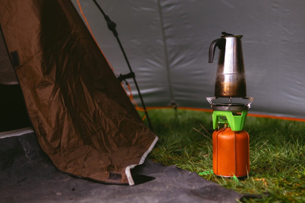 A compact gas burner, a fuel cylinder covered with a brown leather cover, geyser coffee maker. Cooking in the field. Portable camping, tourist equipment. Spring day. Green grass. Tent. Selective focus