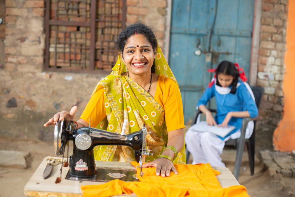 Portrait of happy traditional indian woman wearing sari using sewing machine while her young daughter studying behind her in school uniform.