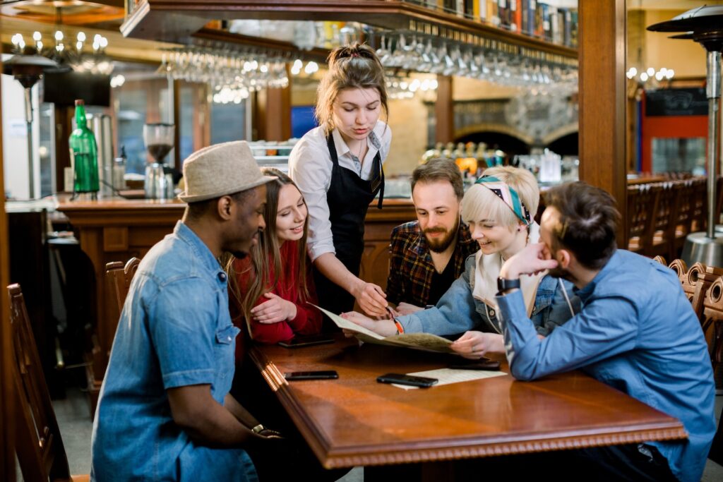 Group of diverse young friends sitting together at a table in a trendy fast food restaurant ordering food from a waitress