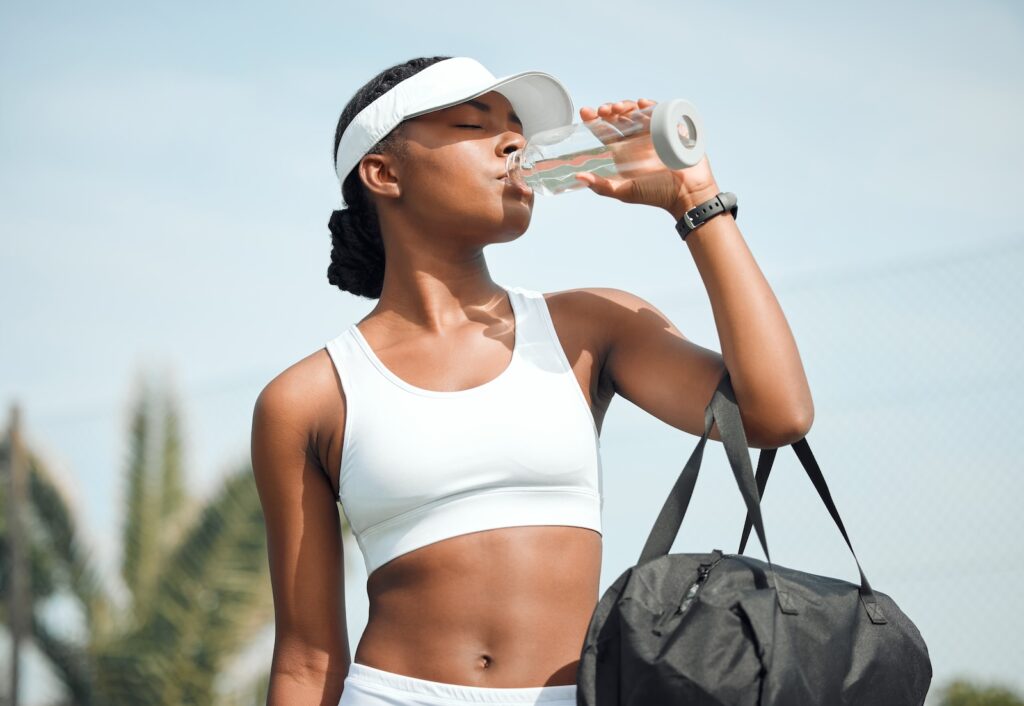 Tennis, drinking water and black woman on court, sports and fitness with nutrition, wellness and workout. African person, outdoor and athlete with bottle racket and training with break and healthy