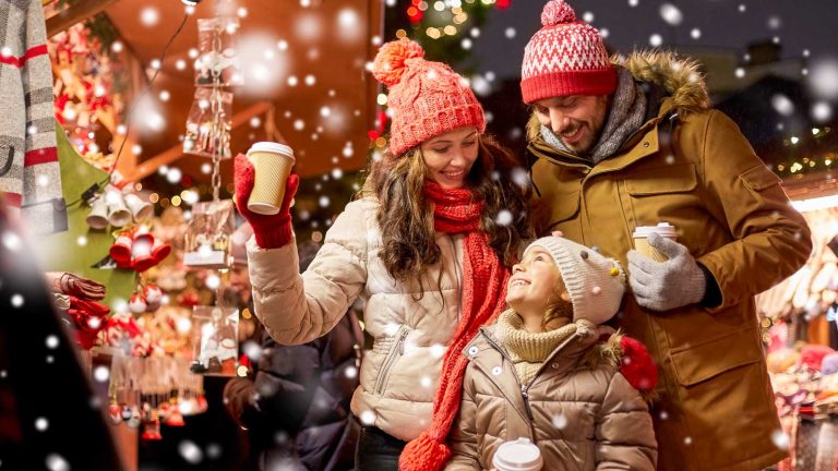 family, winter holidays and celebration concept - happy mother, father and little daughter with takeaway drinks at christmas market on town hall square in tallinn, estonia over snow